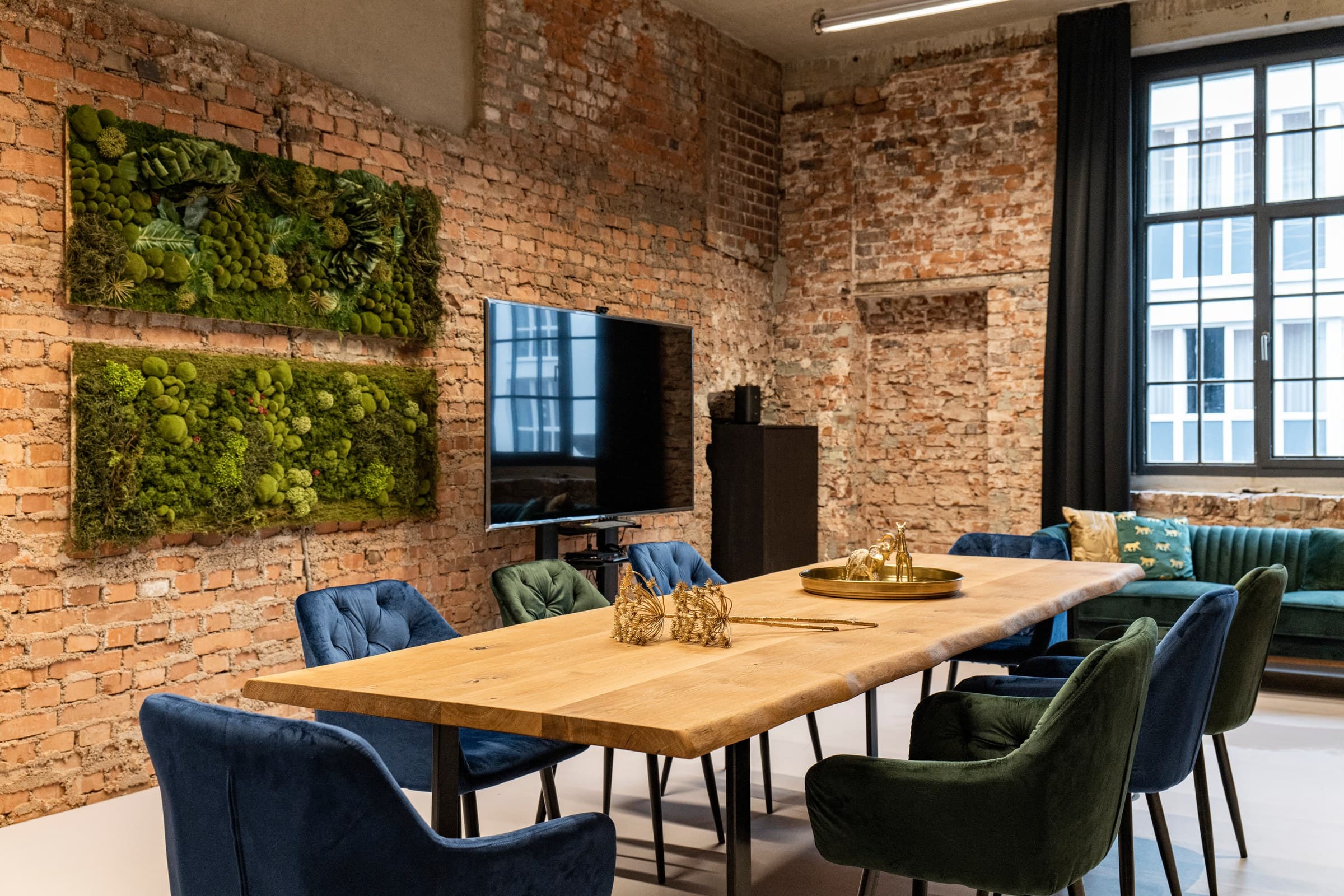 Der Konferenztisch von Werkraum.02 mit blauen und grünen Samtstühlen, an der Backsteinwand zwei Moosbilder und ein großer Bildschirm auf einem Ständer, rechts ein grünes Samtsofa vor einem hohen Sprossenfenster