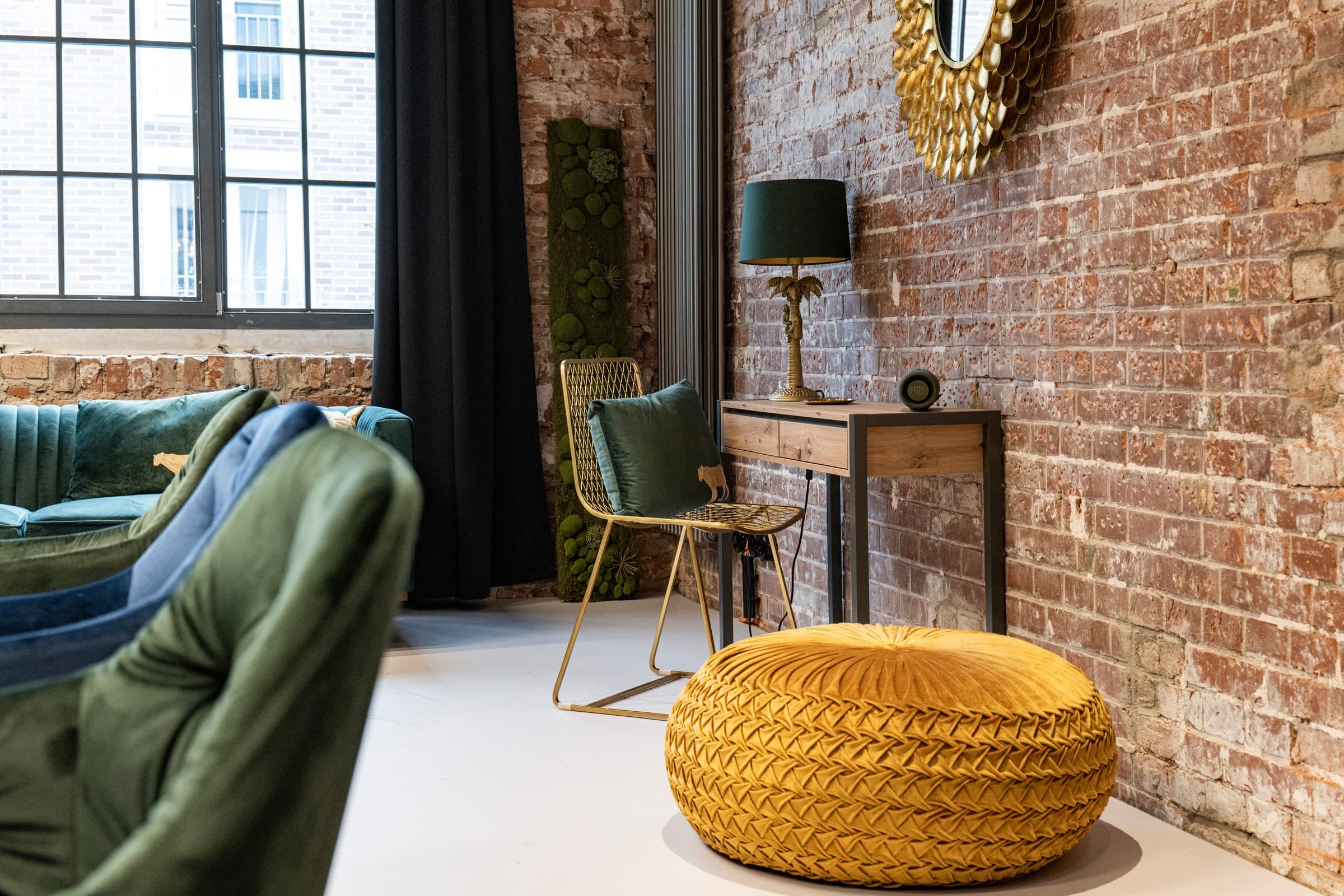 Die Sitzecke von Werkraum.02: ein goldener Drahtstuhl, eine Konsole mit Tischlampe und ein senffarbener Samtpouf vor der Backsteinwand, links ein grünes Samtsofa unter dem Sprossenfenster, vorn angeschnitten zwei Samtstühle