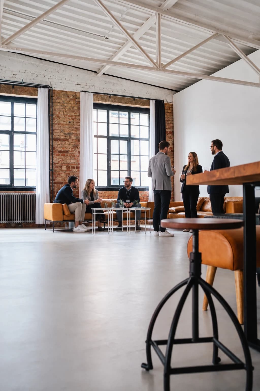 Die Lounge von Werkraum.01 im Gebrauch: drei Gäste auf dem cognacfarbenen Ledersofa, drei weitere stehen mit Gläsern beisammen, vorn ein Hocker am Holztisch