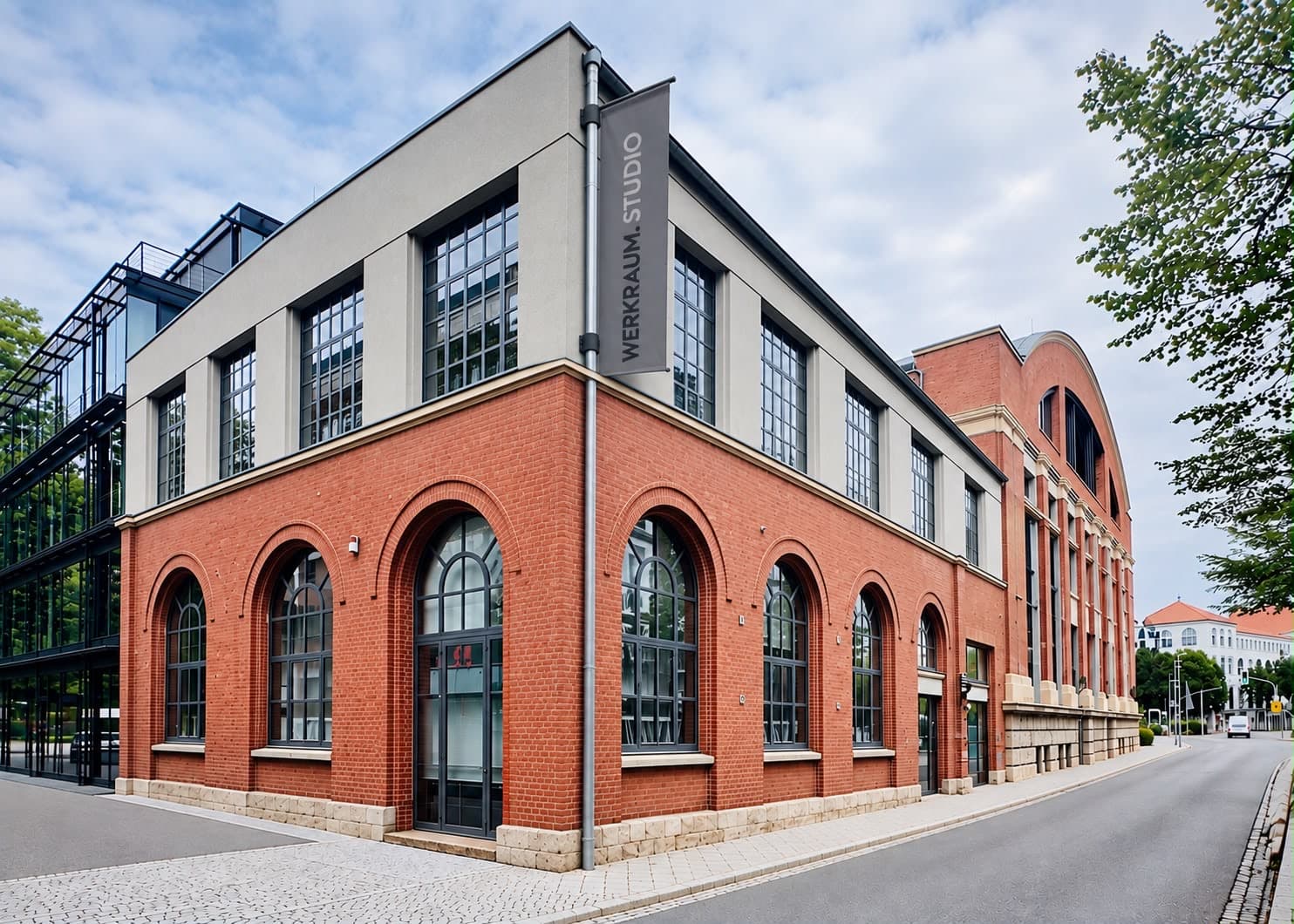 Das Haus über Eck: im Erdgeschoss aus Backstein hohe Rundbogenfenster, im grau verputzten Obergeschoss Sprossenfenster, an der Ecke ein Banner mit der Aufschrift „WERKRAUM.STUDIO“