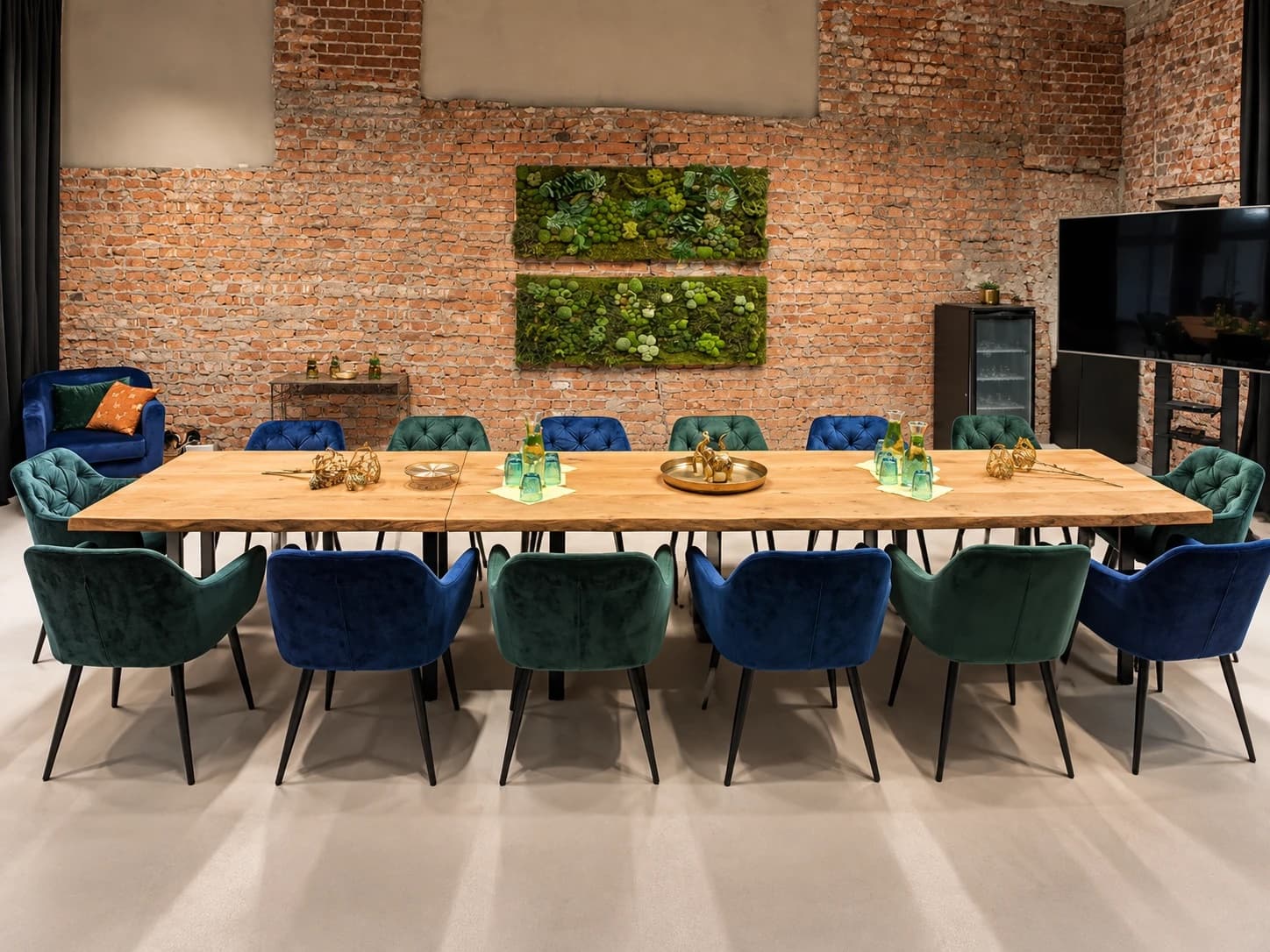 Der lange Konferenztisch von Werkraum.02 mit blauen und grünen Samtstühlen an beiden Längsseiten und an den Stirnseiten, dahinter zwei Moosbilder an der Backsteinwand, links ein blauer Samtsessel, rechts der Bildschirm auf seinem Ständer