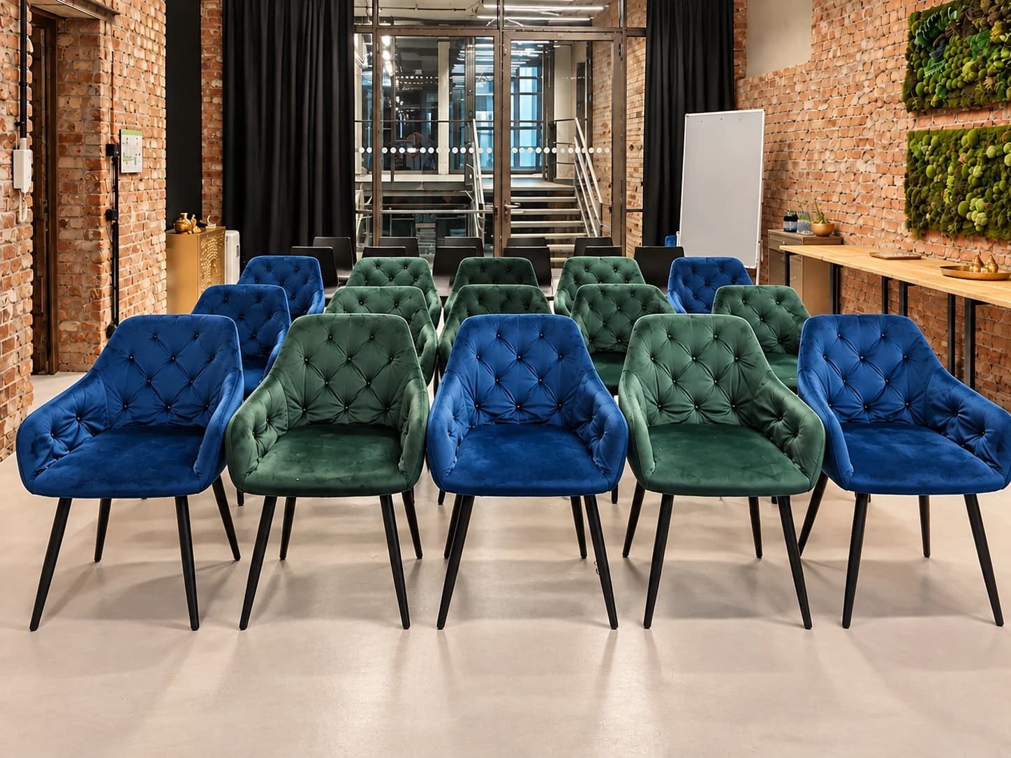 Werkraum.02 in Reihen bestuhlt: blaue und grüne Samtstühle, dahinter schwarze Stühle, vor der Glaswand zum Treppenhaus und einer weißen Stellwand, rechts der beiseitegestellte Konferenztisch unter zwei Moosbildern an der Backsteinwand