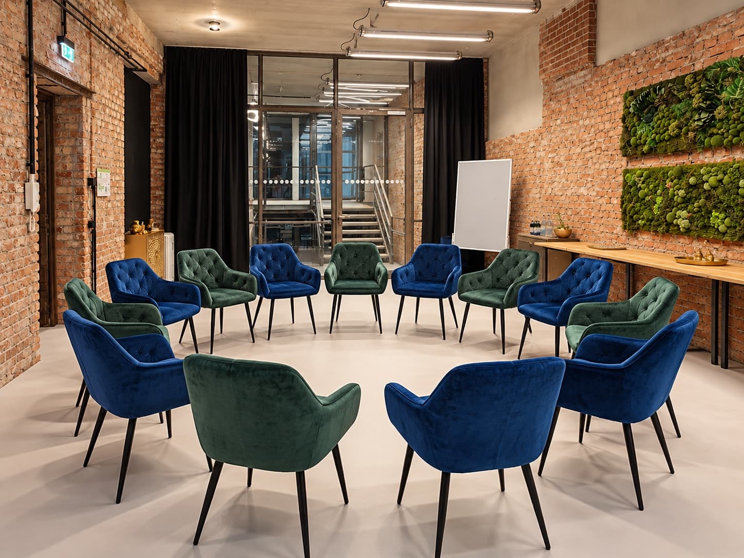 Werkraum.02 mit einem Stuhlkreis aus blauen und grünen Samtstühlen, dahinter die Glaswand zum Treppenhaus und eine weiße Stellwand, rechts der beiseitegestellte Konferenztisch unter zwei Moosbildern an der Backsteinwand