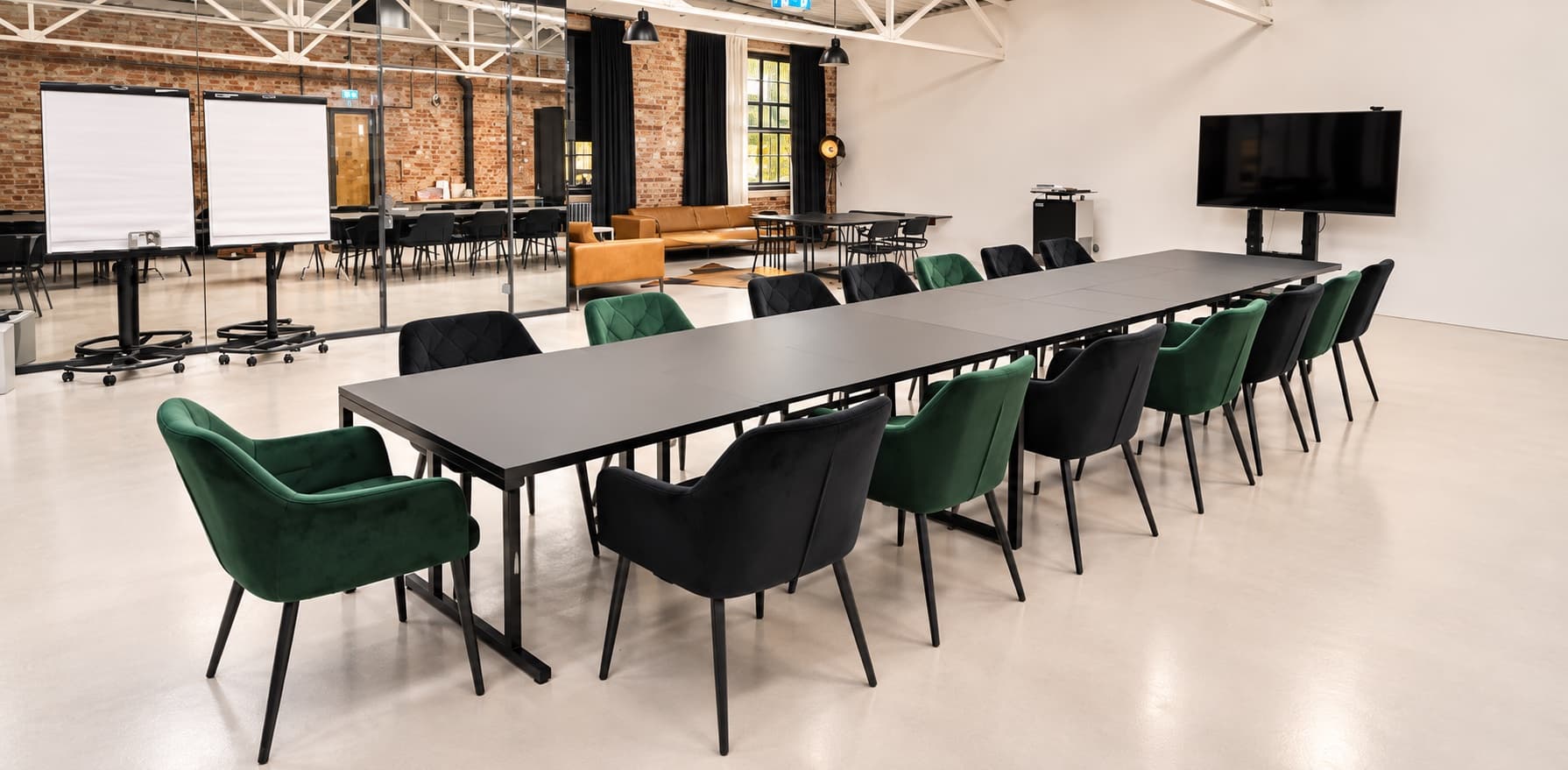 Werkraum.01 mit schwarzen Tischen zu einem langen Block, grüne und schwarze Samtstühle an beiden Längsseiten; links Flipcharts vor der Glaswand eines Nebenraums, hinten die Lounge mit braunen Ledersofas, rechts die weiße Wand