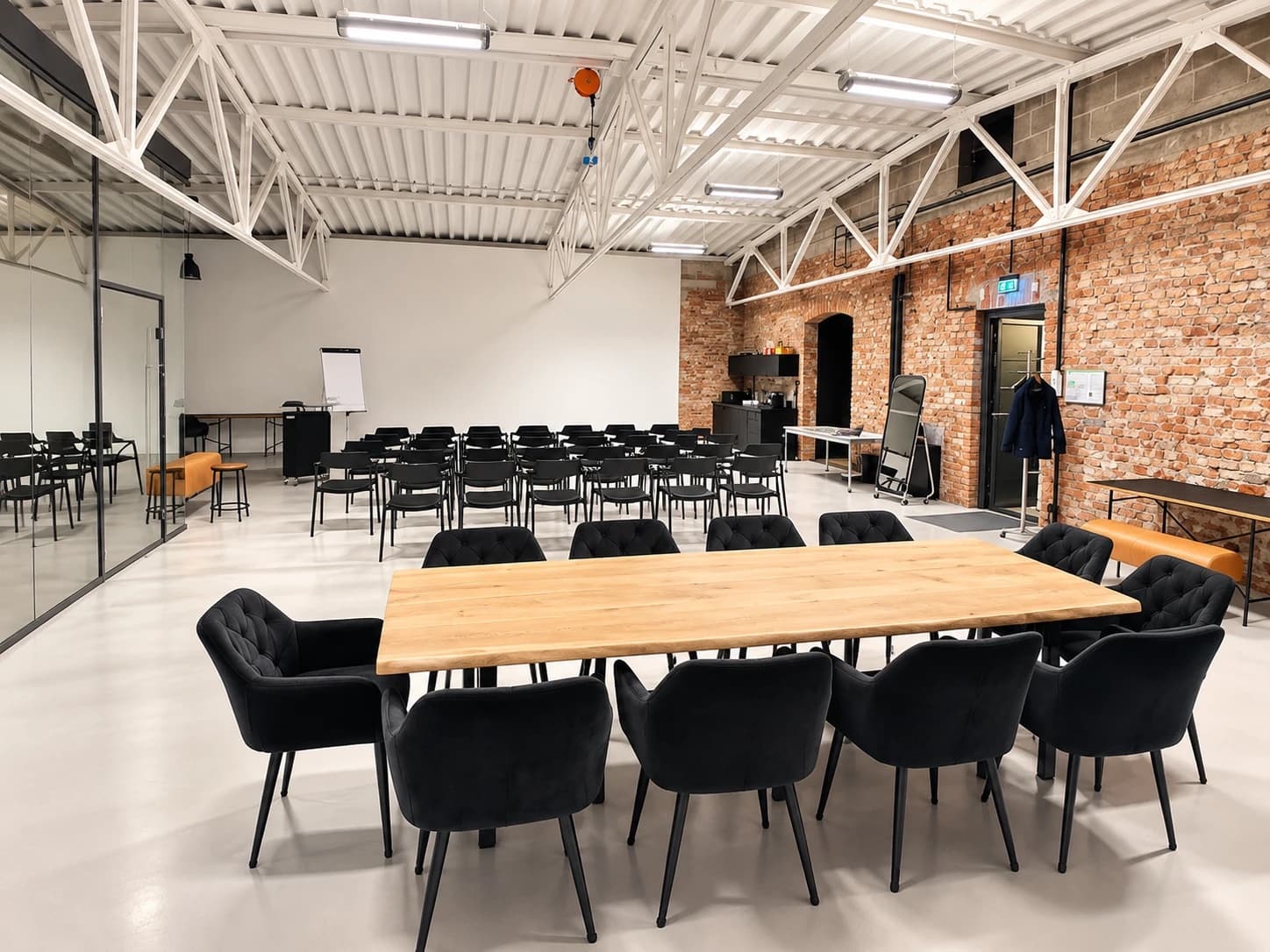 Werkraum.01 individuell gestellt: vorn ein langer Eichentisch mit schwarzen Samtstühlen, dahinter schwarze Stühle in Reihen vor der weißen Wand mit Flipchart, rechts die Backsteinwand mit Küchenzeile und Tür, links die Glaswand eines Nebenraums
