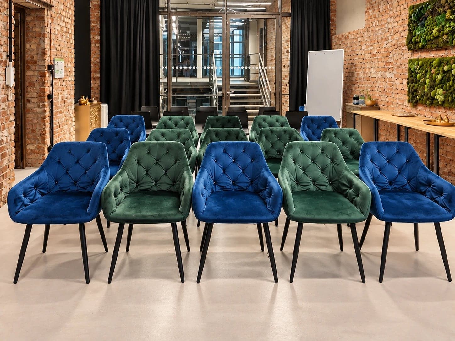 Werkraum.02 in Reihen bestuhlt: blaue und grüne Samtstühle, dahinter schwarze Stühle, vor der Glaswand zum Treppenhaus und einer weißen Stellwand, rechts der beiseitegestellte Konferenztisch unter zwei Moosbildern an der Backsteinwand